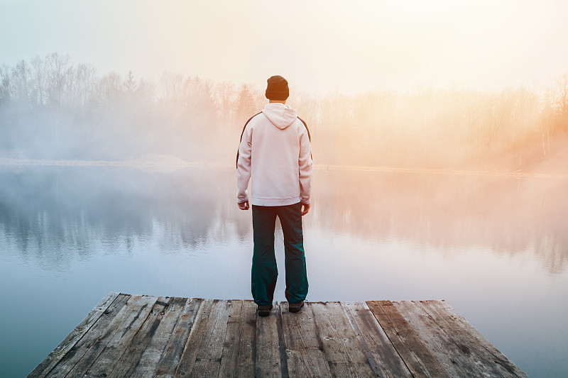 年輕男子在帽衫，帽子和褲子站在木碼頭在池塘岸邊與憂郁的霧日出。捷克晨景圖片下載