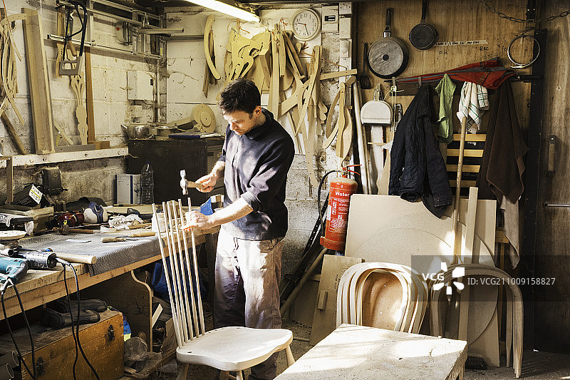 一名男子站在木工车间里，背靠一把木椅干活。图片素材