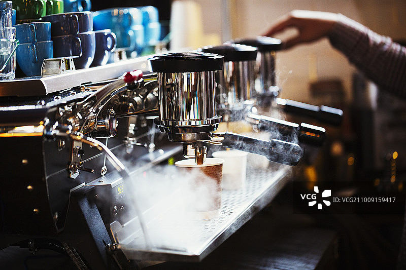 专业咖啡店。一个在大型咖啡机旁工作的人，有三个过滤性的容器、把手和一根散发蒸汽的管道。图片素材