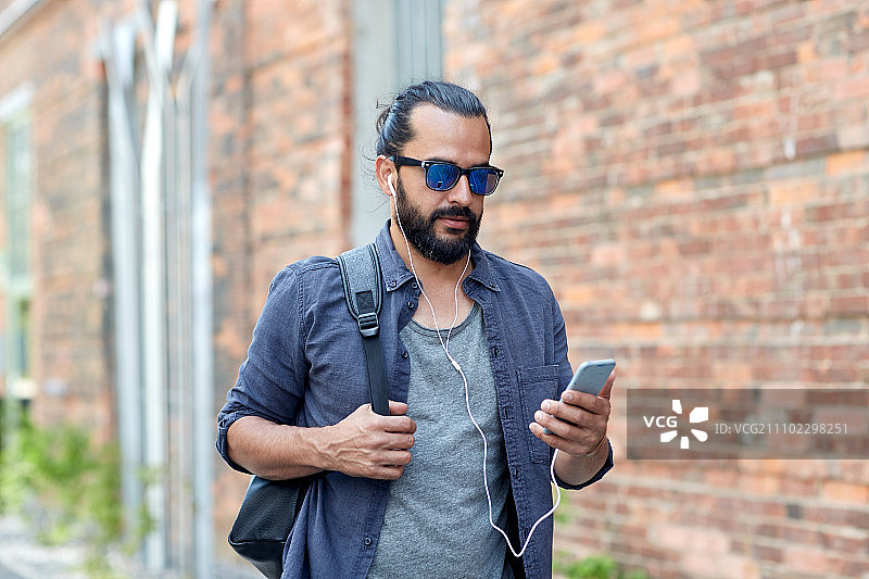 人、技术、旅游和旅游——男人带着耳机、智能手机和包走在城市的街道上，听着音乐图片素材