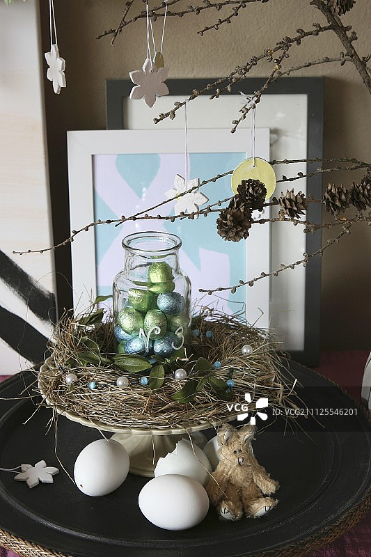 复活节的安排;蛋糕架上放着干草做成的花环和一罐巧克力蛋，下面的桌子上放着蛋壳和毛绒复活节兔子图片素材