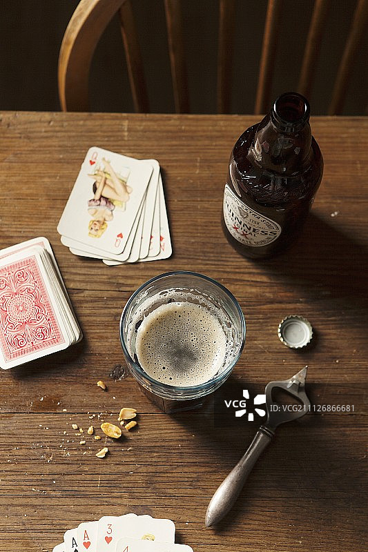 一瓶吉尼斯黑啤酒倒进一个大玻璃杯，放在一张木桌上，周围有瓶盖、金属开瓶器、坚果和扑克牌图片素材