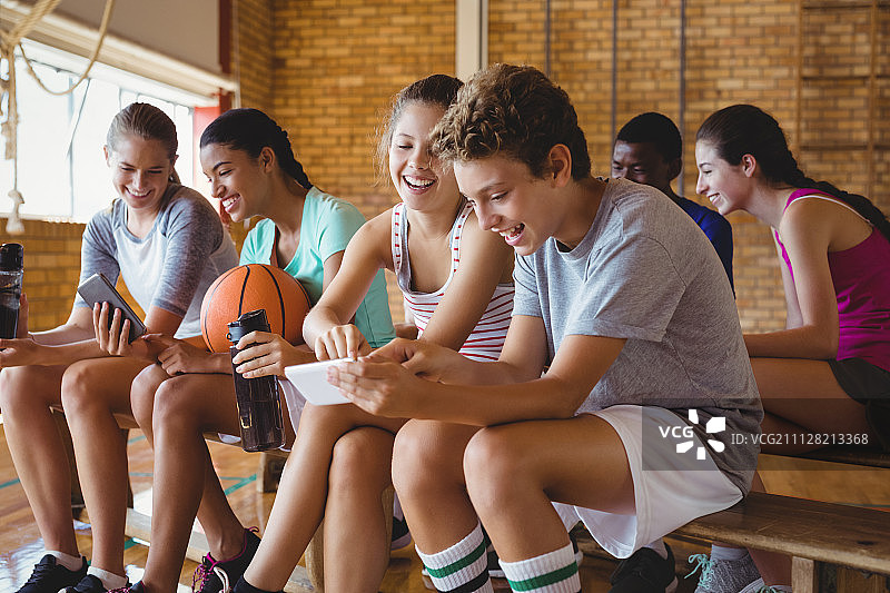 微笑的高中生使用手机，同时放松在篮球场上图片素材