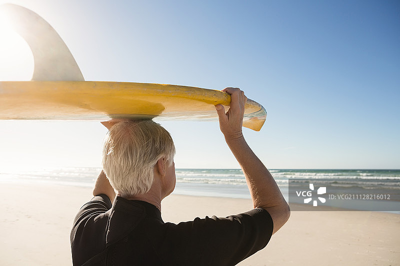 在阳光明媚的日子里，在海滩上背着冲浪板的老人的后视图图片素材