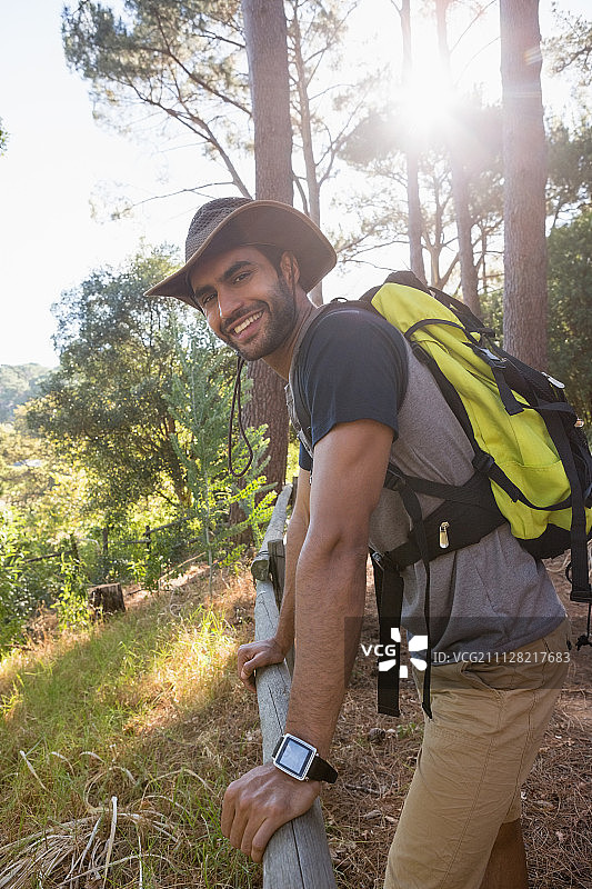 一个背着双肩包的微笑男子倚靠在森林的木栅栏上图片素材
