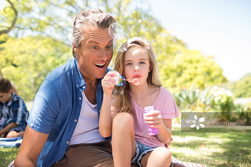 快乐的爸爸和女儿在公园野餐时用泡泡棒吹泡泡图片素材