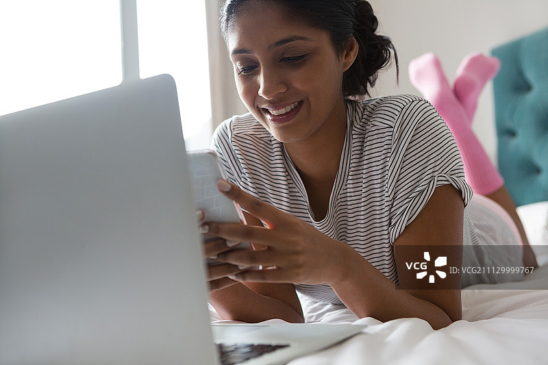 微笑的年轻女子与笔记本电脑使用电话在床上在家图片素材