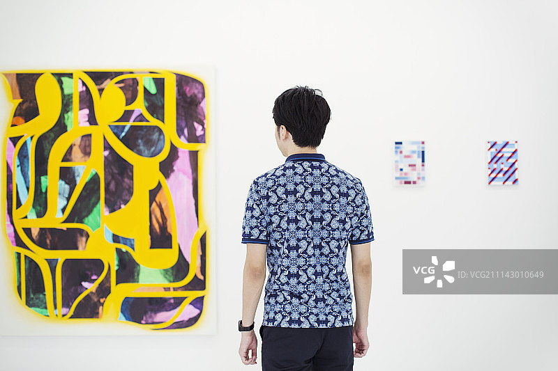 一个穿着蓝色衬衫的黑色短发男子的背影站在画廊里，看着抽象的现代绘画。图片素材