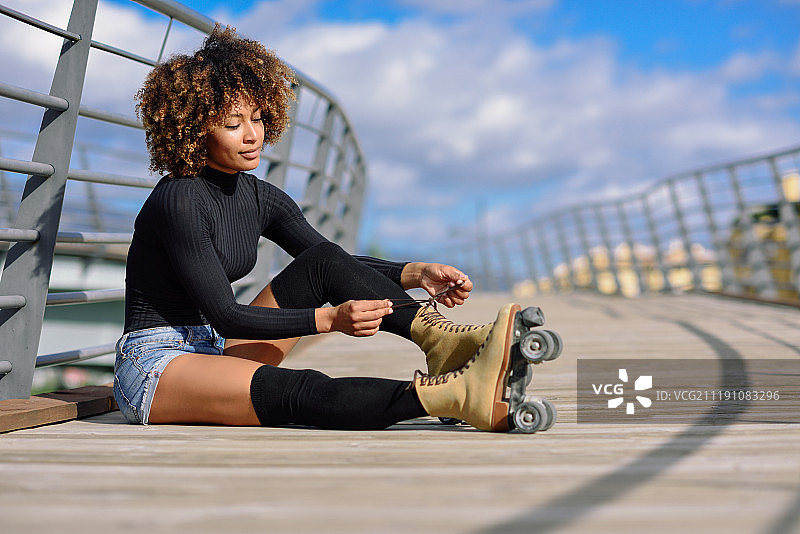 年轻微笑的黑人女孩坐在城市的桥上，穿上溜冰鞋图片素材