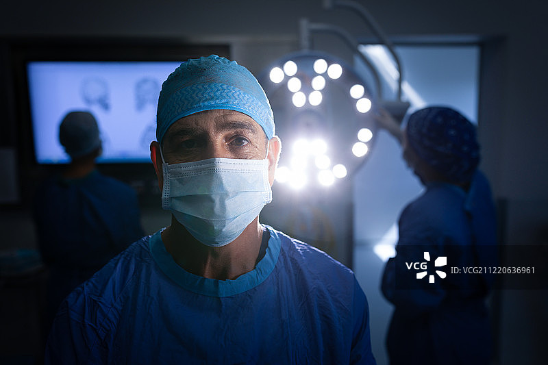 一个白人男性外科医生的肖像，戴着手术面具，看着摄像机在手术室和后面的同事工作图片素材