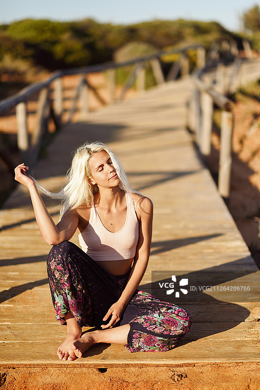 在西班牙安达卢西亚的加的斯，一名女子在美丽的海滩上欣赏日落。年轻女子微笑着坐在木桥上微笑的女人在美丽的海滩上欣赏日落图片素材