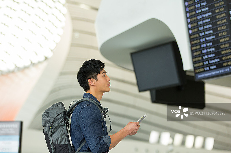 青年男子背着包拿着护照和机票在机场看显示屏核对飞机航班信息图片素材