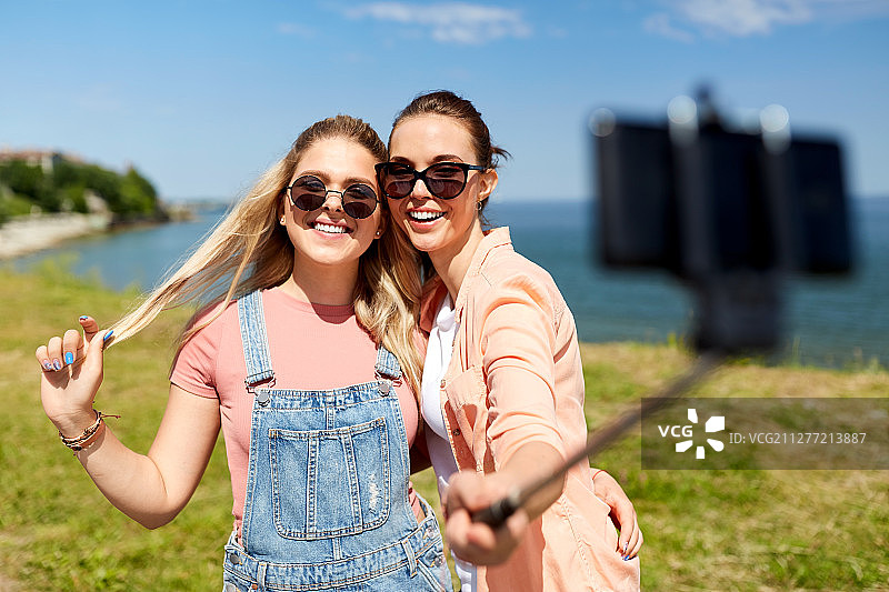 休闲和友谊的概念——快乐微笑的少女或朋友们戴着太阳镜拥抱，在夏天的海边用智能手机自拍。少女或朋友在夏天自拍图片素材