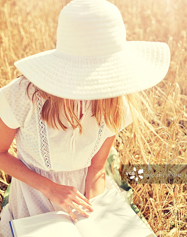 国家，暑假，文学和人的概念-近距离的年轻女子在白色草帽和服装阅读在谷物田。近距离的女人阅读谷物田的书图片素材