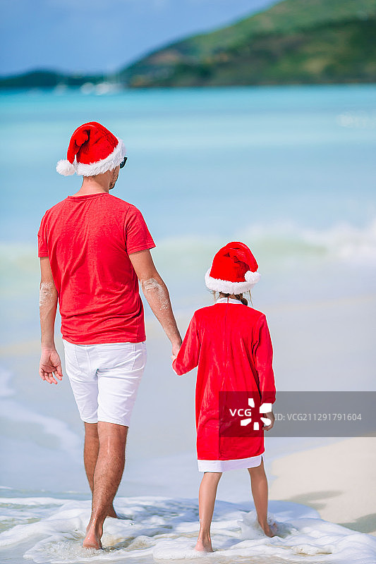 父亲和女儿戴着圣诞老人帽在热带海滩上散步图片素材