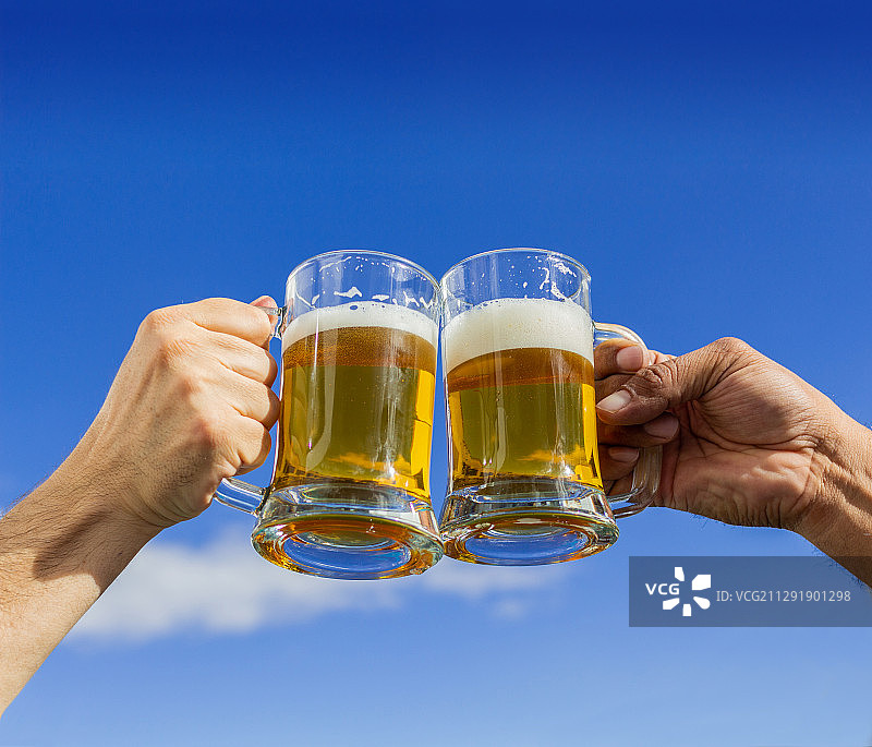 一对举起啤酒杯的手臂，在蓝天的背景下庆祝图片素材