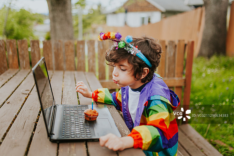 加拿大维多利亚，一个穿着彩虹夹克、坐在笔记本电脑上的小男孩在纸杯蛋糕上许下生日愿望图片素材