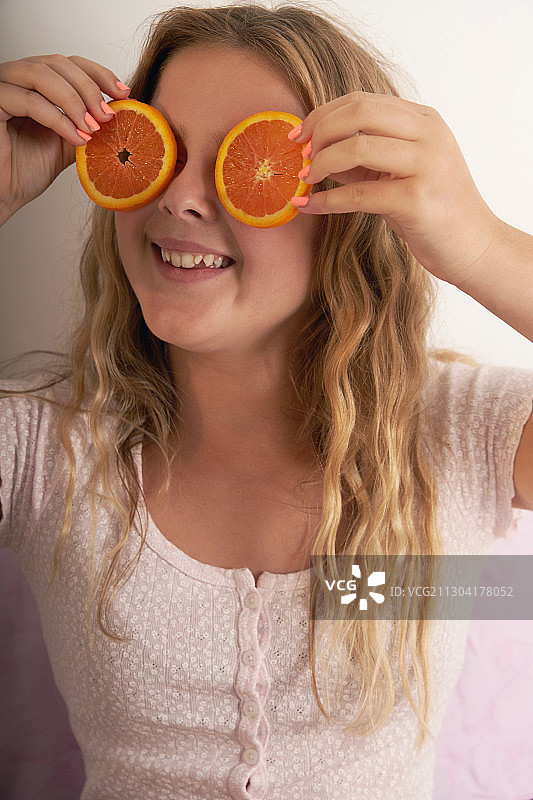 快乐的年轻女孩拿着橙色的水果片在她的眼睛前面，在白色的背景图片素材