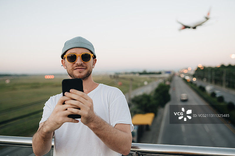 泰国清迈，一名戴着帽子和太阳镜的白人男子站在桥上使用智能手机图片素材
