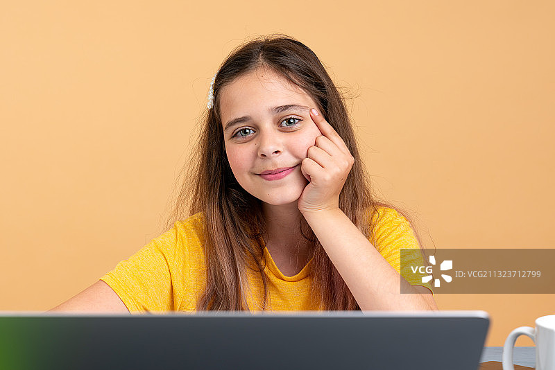 微笑的女孩的肖像与笔记本电脑坐在桌子上，以彩色的背景图片素材
