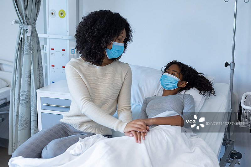 混血母亲在病床上安抚生病的女儿，手牵着手，两人都戴着口罩图片素材