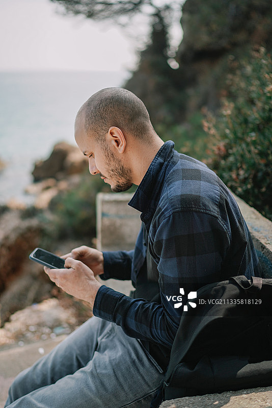 西班牙海边，一名男子坐在岩石上玩手机图片素材