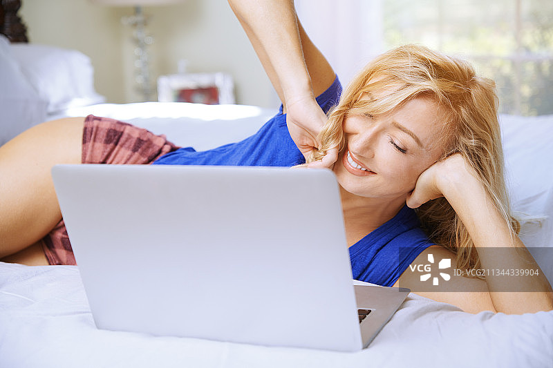 美丽的金发自由职业女人躺在床上，使用笔记本电脑图片素材