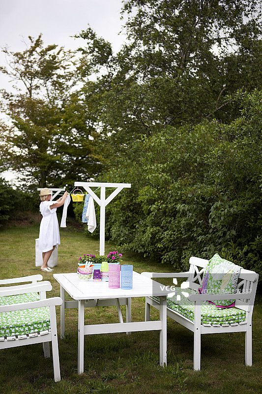 桌子和椅子由白色的漆木制成，花园中有绿色和白色的坐垫，背景中是坐在晾衣架上的女人图片素材
