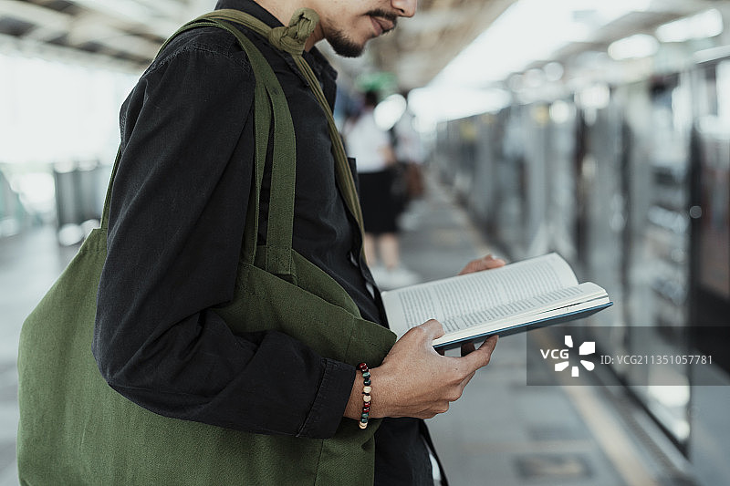 一个男人在轻轨站等火车时，手里拿着一本打开的书。图片素材