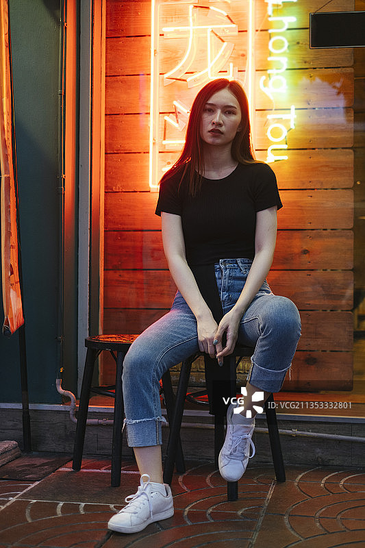 时尚肖像-美丽的时髦亚洲年轻女子在黑色衬衫和牛仔裤裤子坐在椅子上霓虹灯背景。图片素材