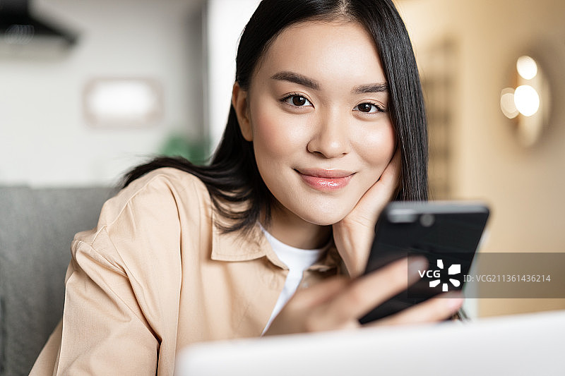 微笑的亚洲女性在家里用笔记本电脑工作，看着智能手机图片素材