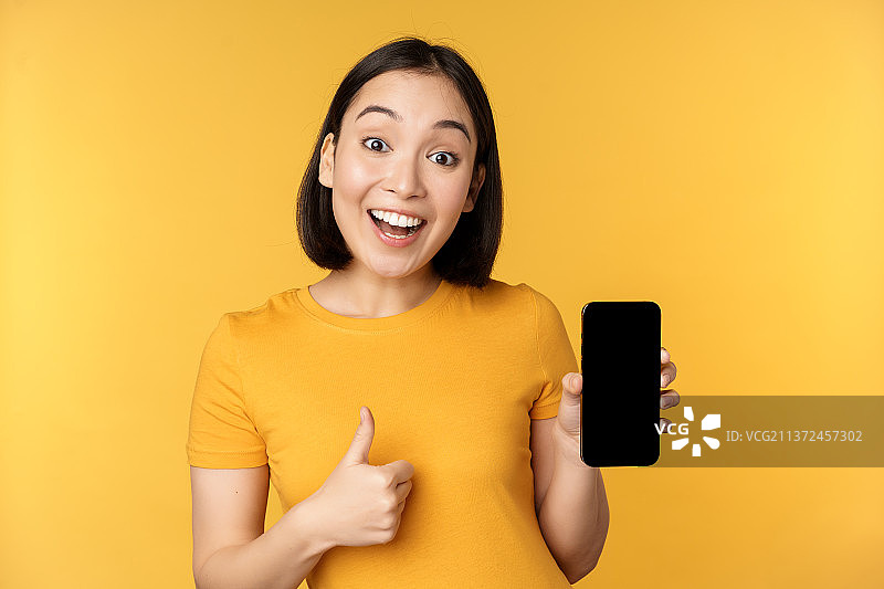 兴奋的亚洲女人展示手机屏幕，竖起大拇指，像史密斯一样图片素材