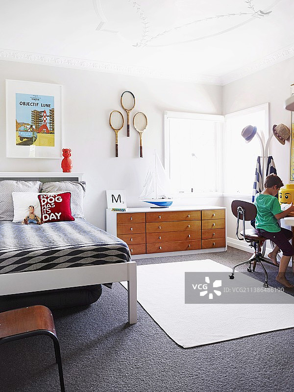 孩子的卧室——男孩坐在椅子上，旁边是衣架，床，餐具柜和挂在墙上的网球拍图片素材