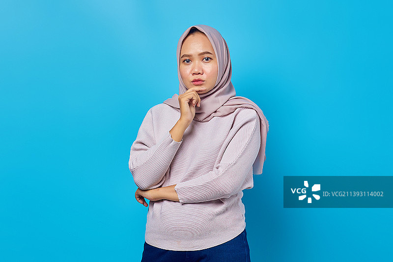 有吸引力的亚洲女人的肖像思考和触摸下巴的蓝色背景，印度尼西亚图片素材