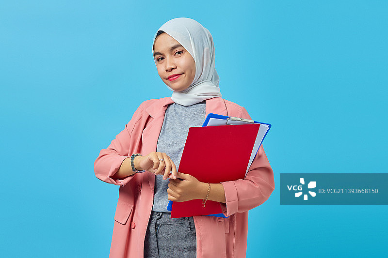 肖像开朗的亚洲妇女拿着文件夹和看手表的蓝色背景，印度尼西亚图片素材