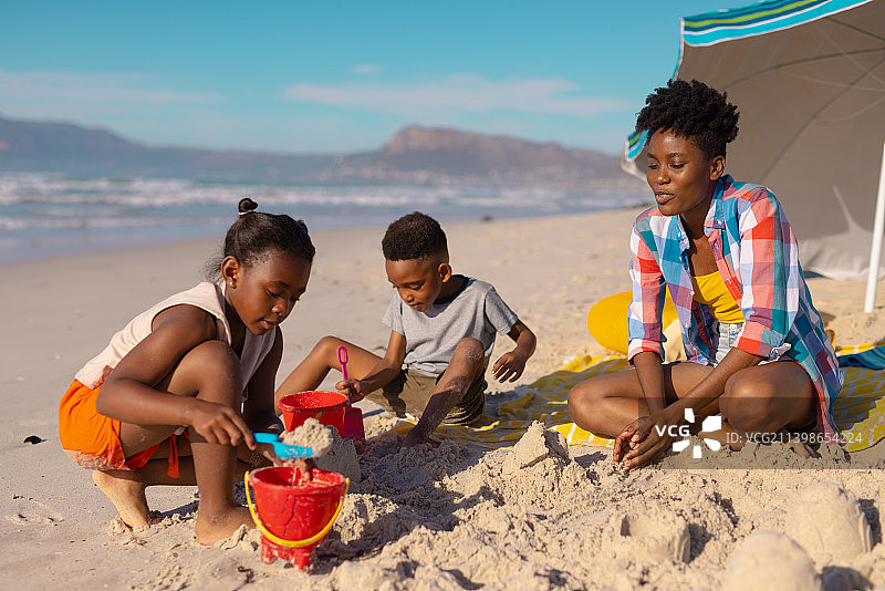 非洲裔美国年轻母亲与儿子和女儿坐在海滩上玩沙子，对着天空图片素材