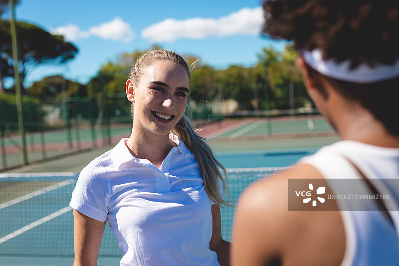 在阳光灿烂的日子里，微笑的白人女运动员在网球场上看着非洲裔美国运动员图片素材