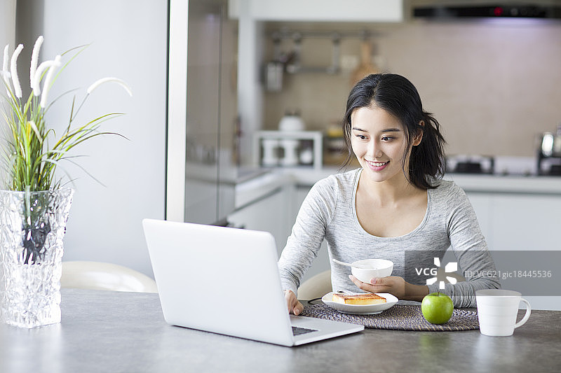 年轻女子一边吃早餐一边使用笔记本电脑图片素材