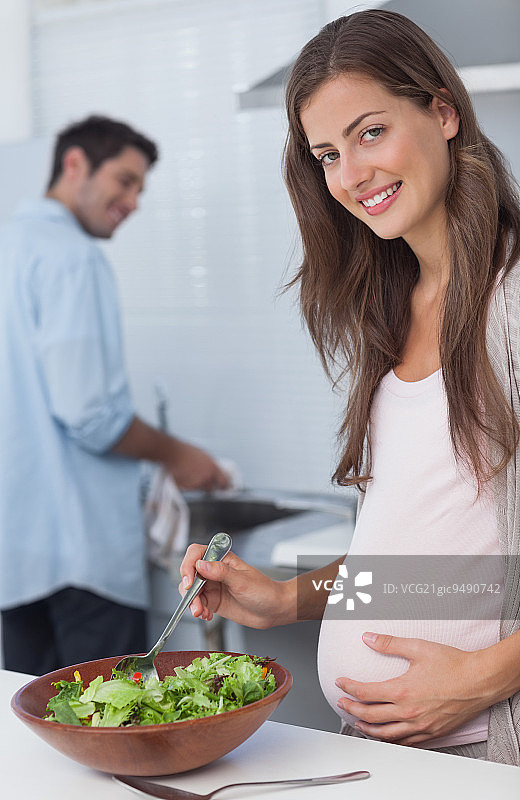 一名孕妇在厨房准备沙拉，她的丈夫在后面洗碗图片素材