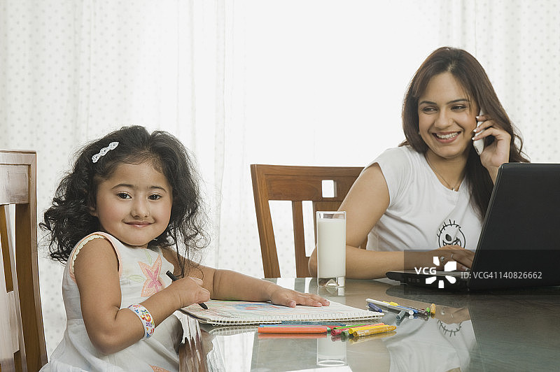 小女孩在餐桌旁用笔记本电脑给一幅画上色，她的妈妈也在用图片素材