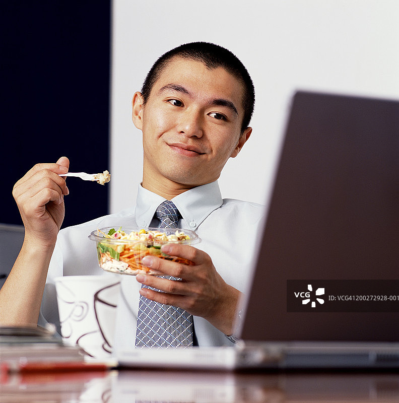 年轻的商人坐在办公桌前吃午饭，看着笔记本电脑图片素材