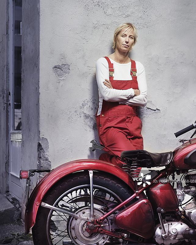 一个成熟的女人靠在摩托车旁，双臂交叉，肖像图片素材