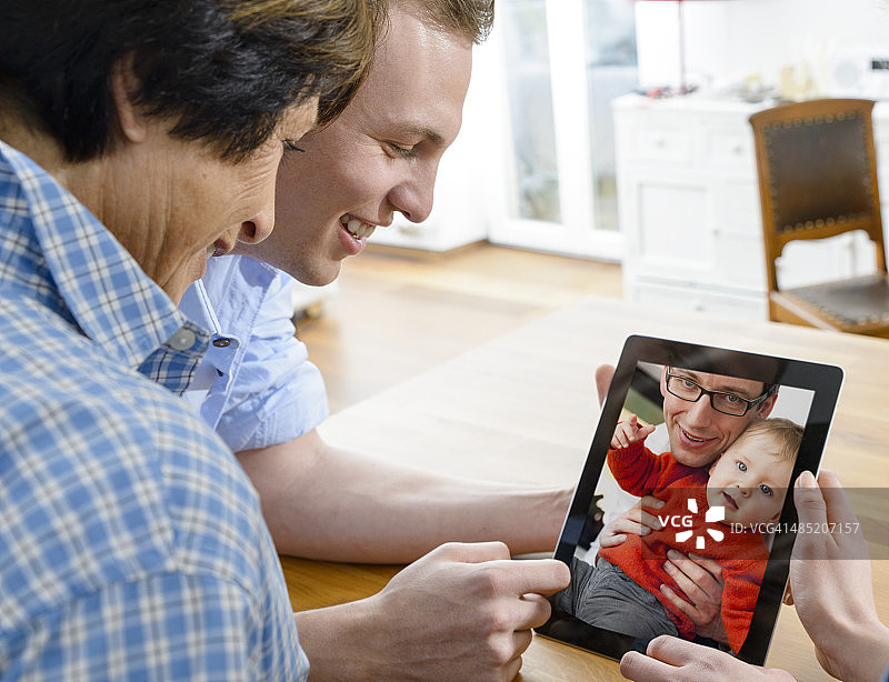 年长的妇女和年轻的男子看着图像的父亲和婴儿的儿子在数码平板电脑图片素材