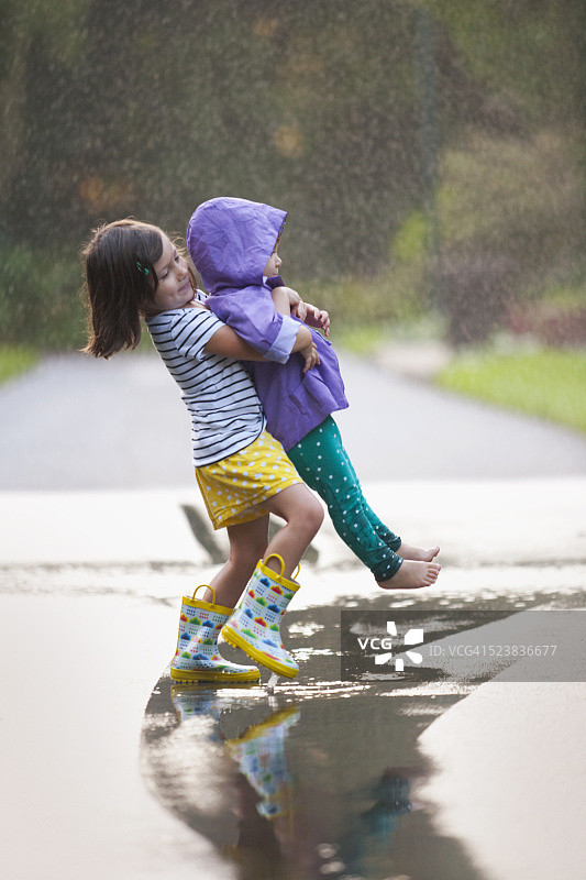 女孩抱着蹒跚学步的妹妹穿过街道上的水坑图片素材
