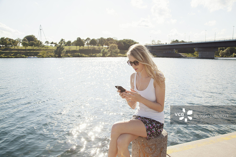 奥地利维也纳多瑙河岛，一名年轻女子坐在河边用智能手机发短信图片素材