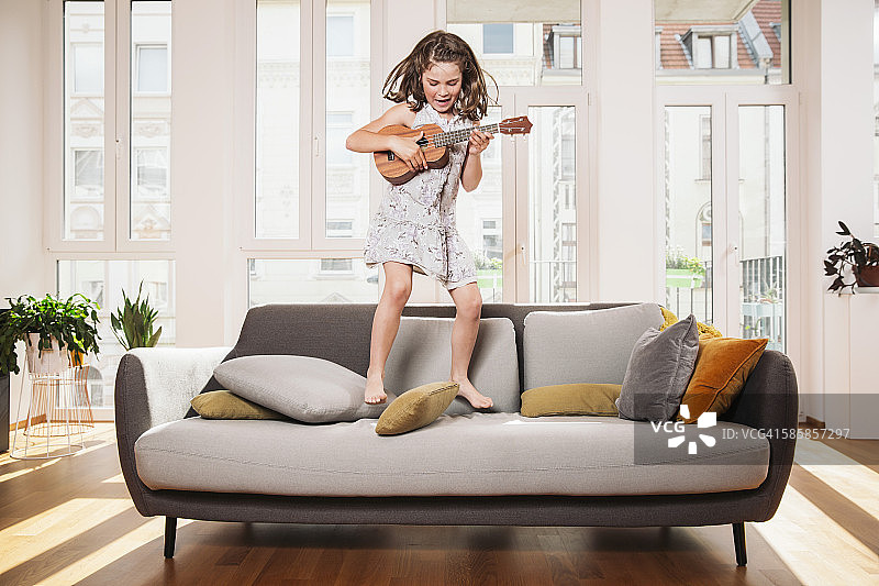 快乐的女孩玩迷你吉他，同时跳在家里客厅的沙发上图片素材