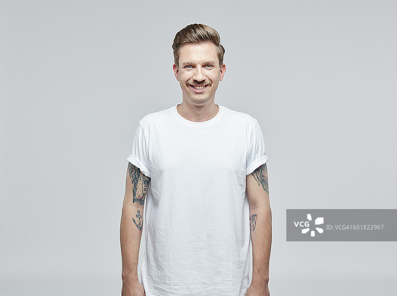 肖像的微笑男子与纹身在他的手臂上穿着白色t恤在前面的灰色背景图片素材