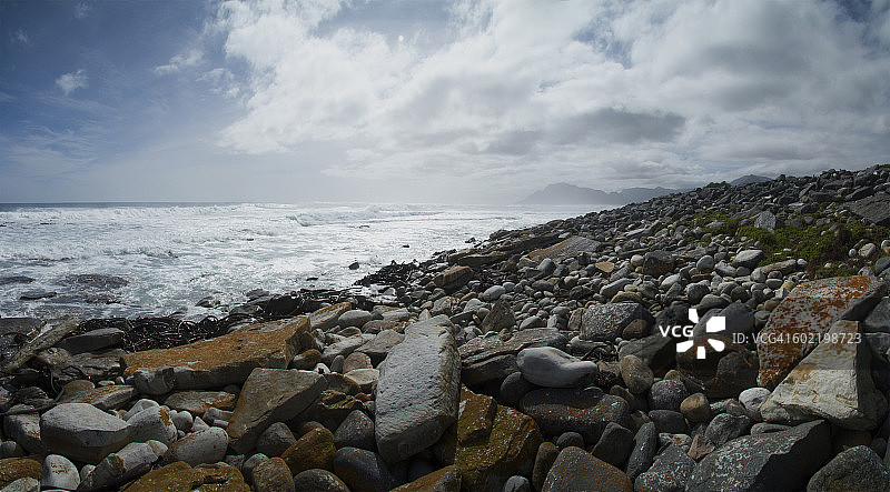岩石遍布的海滩和海浪下一个戏剧性的多云的天空，豪特湾在远处，Slangkoppunt, Kommetjie，开普敦，西开普，南非。图片素材