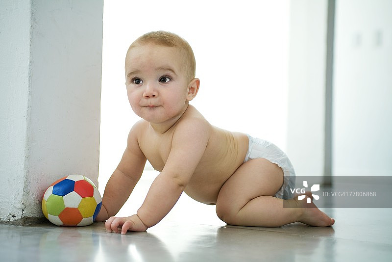 婴儿在地板上爬行，靠近球，看向别处，咬着嘴唇图片素材