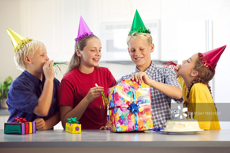 朋友们带着帽子和噪音制造者聚集在一起，一个男孩打开了生日礼物图片素材